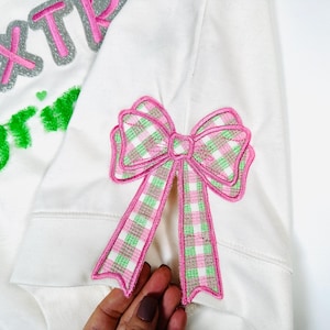 May include: White sweatshirt with pink and green embroidered bow on the sleeve. The text "EXTREME" is embroidered in silver and pink on the front of the sweatshirt.