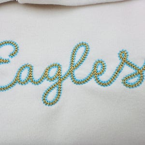 Puede incluir: Primer plano de una tela blanca con la palabra "Eagles" bordada en letra cursiva. El bordado presenta un hilo dorado delineado con un hilo azul claro, creando un efecto texturizado.