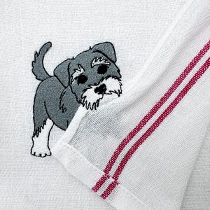 Könnte beinhalten: Weißes Küchentuch mit einem gestickten Schnauzer-Hund-Design. Der Hund ist grau und weiß mit schwarzen Augen und einer schwarzen Nase. Das Handtuch hat rote Streifen am Rand.