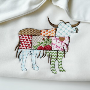 May include: A white fabric baby blanket with a colorful patchwork design of a highland cow embroidered on it.