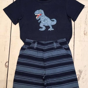 K&ouml;nnte beinhalten: Ein blaues T-Shirt mit kurzen &Auml;rmeln und einem blauen und grauen Dinosaurier-Motiv. Das Shirt wird mit einer blauen und grauen gestreiften Hose kombiniert.