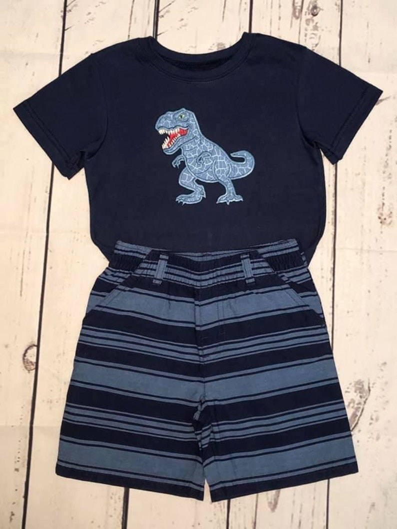 K&ouml;nnte beinhalten: Ein blaues T-Shirt mit kurzen &Auml;rmeln und einem blauen und grauen Dinosaurier-Motiv. Das Shirt wird mit einer blauen und grauen gestreiften Hose kombiniert.