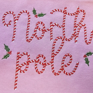 May include: Pink sweatshirt with "North Pole" embroidered in red and white candy cane stripes with green holly leaves.
