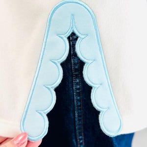 May include: A white garment featuring a light blue scalloped applique detail along a vertical slit. The applique has a smooth, rounded edge design with visible stitching. Beneath the slit, a dark blue denim fabric is visible.