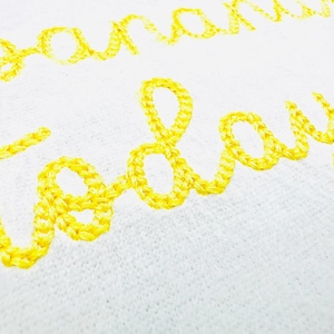 May include: Close-up of white fabric with yellow embroidered text that reads "Banana today". The text is in a cursive style, and the embroidery has a textured, raised appearance. The background is a plain white.