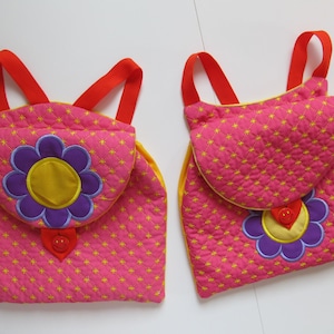 以下が含まれることがあります： 黄色い縁取りと刺繍された花の模様が施された、ピンク色のキルティングのバックパックが2つ。それぞれのバックパックには、笑顔の顔をした黄色のボタンが付いています。