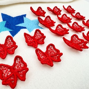 May include: Close-up of red lace bows and blue embroidered stars on a white background. The bows are arranged in a scattered pattern, while the stars are grouped together. The bows and stars are likely for crafting or embellishment.