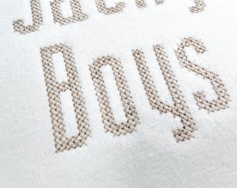 May include: Close-up of a white fabric with the embroidered text "JACK'S BOYS" in a light brown color. The letters are large and blocky, with a cross-stitch design.