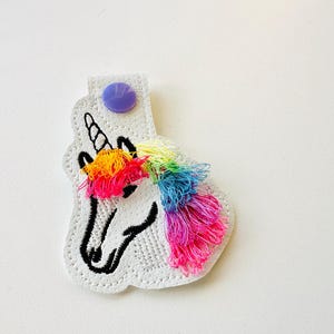 Op de afbeelding: Een witte eenhoornvormige sleutelhanger met een regenboogmanen en een paarse knoop. De eenhoorn heeft een zwarte omtrek en een gestikt ontwerp. De manen zijn gemaakt van kleurrijke draden in tinten geel, oranje, roze en blauw.