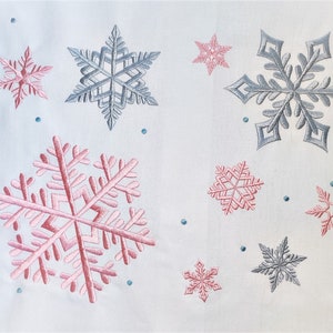 May include: A white background with nine embroidered snowflakes. Six of the snowflakes are pink and three are blue.