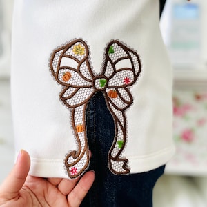 May include: White top with a large, embroidered bow design. The bow features a brown outline with a lace-like pattern and is decorated with small pumpkins, leaves, and flowers in fall colors. The top is paired with dark blue jeans.