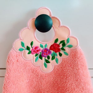 May include: A pink hand towel with a scalloped edge and embroidered flowers in shades of pink, purple, and green. The towel has a loop for hanging.
