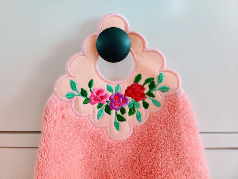 May include: A pink hand towel with a scalloped edge and embroidered flowers in shades of pink, purple, and green. The towel has a loop for hanging.