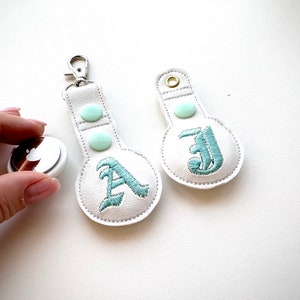以下が含まれることがあります： 薄い青色のボタンと刺繍された文字が付いた白い革のキーホルダーが2つ。左側のキーホルダーには「A」の文字が、右側のキーホルダーには「J」の文字が刺繍されています。