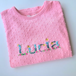 May include: Pink sweatshirt with a floral embroidered name "Lucia" on the front.