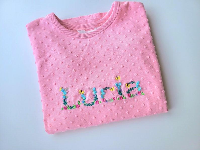 May include: Pink sweatshirt with a floral embroidered name "Lucia" on the front.