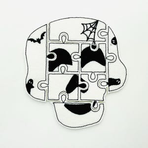May include: A white Halloween-themed puzzle in the shape of a skull, with black accents. The puzzle pieces feature a bat, spiderweb, and ghost. The design is outlined in black stitching.