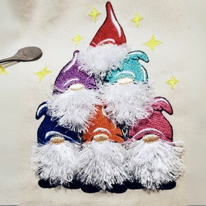 May include: A stack of six embroidered gnomes with white beards and colorful hats. The gnomes are arranged in a pyramid shape with the tallest gnome at the top. The gnomes are all smiling and have a whimsical look.