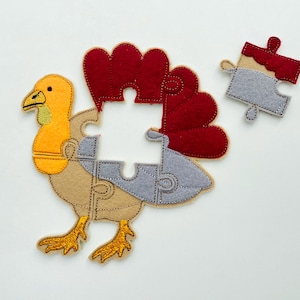 May include: A felt turkey puzzle with pieces in shades of brown, gray, red, and yellow. The turkey's body is tan, with a red wattle and tail feathers. The puzzle is designed for children.