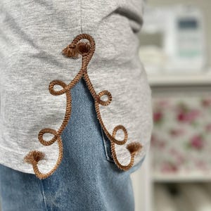 以下が含まれることがあります： 茶色の装飾的なデザインが施されたグレーのTシャツのクローズアップ。 デザインはループとタッセルのシリーズです。 シャツはブルージーンズと組み合わせています。