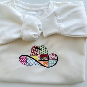 May include: White sweatshirt featuring an embroidered patchwork cowboy hat design in various colours.