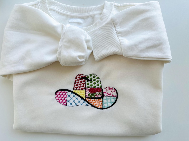 May include: White sweatshirt featuring an embroidered patchwork cowboy hat design in various colours.