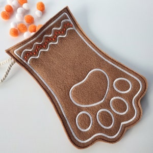 Puede incluir: Una media de fieltro marrón con un diseño de huella de pata blanca y un lazo de cuerda blanco. La media está llena de pompones naranjas y blancos.