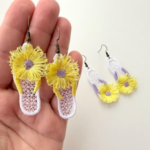 May include: A pair of white crocheted flip-flop earrings with yellow flowers and purple centers. The earrings are hanging from a hand.