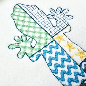 May include: Embroidered design of a whimsical lizard with a patchwork pattern. The lizard features green gingham, blue zigzags, and yellow star accents on a white background. The design includes a detailed outline.
