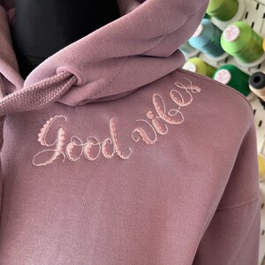 May include: A mauve hoodie with a drawstring hood and the words "Good vibes" embroidered in pink on the front.