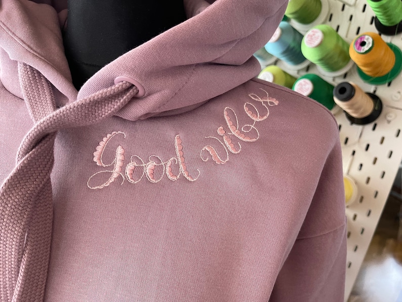 May include: A mauve hoodie with a drawstring hood and the words "Good vibes" embroidered in pink on the front.