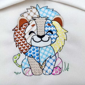 Può includere: Ricamo di un leone con criniera e corpo patchwork in vari colori, tra cui marrone, blu e giallo. Il leone ha una faccia sorridente ed è seduto su uno sfondo bianco con erba ricamata.