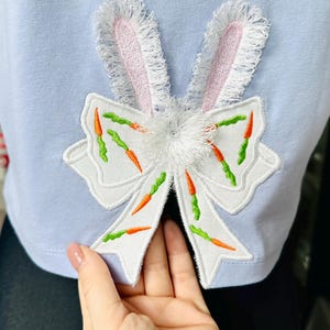 Può includere: Una maglietta blu chiaro con un fiocco bianco ricamato con dettagli di carote e un design a orecchie di coniglio rosa.