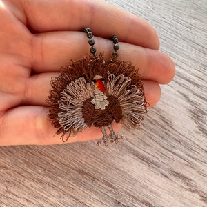 May include: A brown and white embroidered turkey charm on a black chain necklace. The turkey has a red wattle and a brown body with a white tail.
