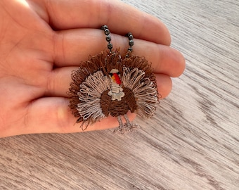May include: A brown and white embroidered turkey charm on a black chain necklace. The turkey has a red wattle and a brown body with a white tail.