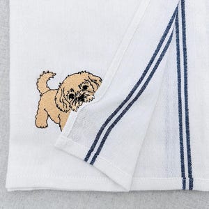 Pode incluir: Toalha de cozinha branca com um cachorrinho bordado bege espreitando do canto. A toalha tem listras azul marinho na borda. A toalha está dobrada, mostrando o desenho do cachorrinho e os detalhes das listras.