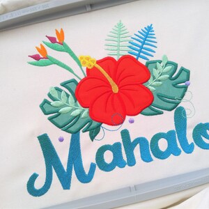 May include: Embroidered design featuring a red hibiscus flower, green leaves, and the word "Mahalo" in blue cursive lettering.