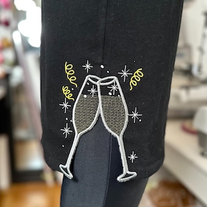 Puede incluir: Camiseta negra con un diseño bordado de dos copas de champán brindando, con confeti amarillo y estrellas blancas.
