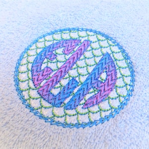 May include: A blue and pink embroidered monogram on a light blue towel. The monogram is surrounded by a green and blue scalloped border.