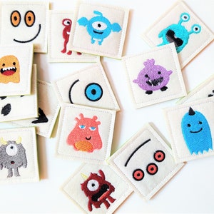 May include: A set of 14 felt squares with embroidered monster faces. Each square features a different monster with various colors, including blue, red, orange, purple, and green. The monsters have different facial expressions, including smiling, frowning, and surprised.