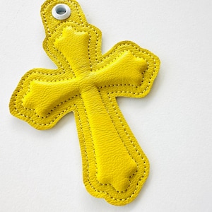 May include: A yellow leather cross with a white button on the top. The cross is stitched with a yellow thread.