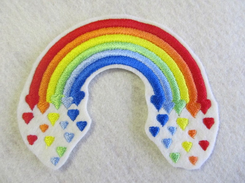May include: A rainbow-colored embroidered patch with a white background. The rainbow is made up of red, orange, yellow, green, blue, and purple. There are small hearts of different colors falling from the rainbow.