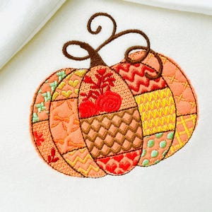 Peut inclure: Citrouille brodée avec des sections aux motifs variés dans les tons orange, jaune et marron. La citrouille a une tige marron et un motif décoratif cousu. Le fond est en tissu blanc.