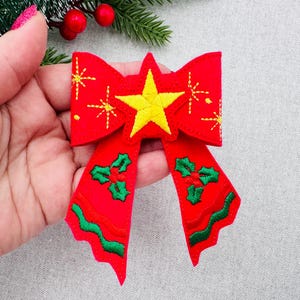 May include: A red Christmas bow with a yellow star in the center and embroidered starbursts. The bow features green holly and a wavy green border on the tails. Festive holiday decor.