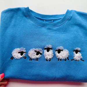 Op de afbeelding: Een lichtblauw sweatshirt met een rij geborduurde witte schapen met zwarte gezichten.