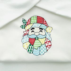 May include: Embroidered Santa Claus face on white fabric. The Santa's hat, beard, and face are made of colourful patchwork patterns. The design includes holly leaves and berries, creating a festive holiday theme.