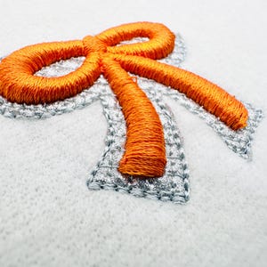 Op de afbeelding: Close-up van een gebordenv oranje strik met een zilveren omtrek op een witte achtergrond. De strik is ingewikkeld gestikt, wat de textuur van de draad laat zien. De zilveren omtrek voegt een subtiele glans toe en creëert een decoratief element.