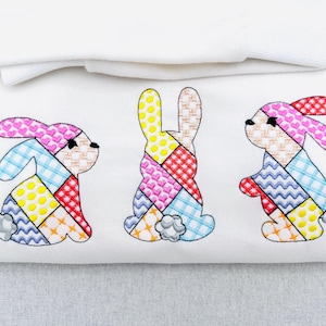 May include: White fabric featuring three embroidered bunnies with patchwork designs. The bunnies have colorful patterns, including hearts, checks, and polka dots. The design is on a white background, with a folded white fabric at the top.