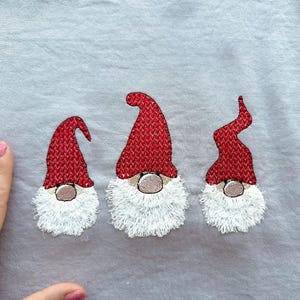 May include: Three embroidered gnomes with red hats and white beards on a light gray fabric background.