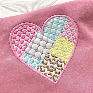 May include: A pink sweatshirt with a large embroidered heart. The heart is made up of different colored patches, including pink, blue, yellow, and brown. The patches are stitched together in a patchwork style.
